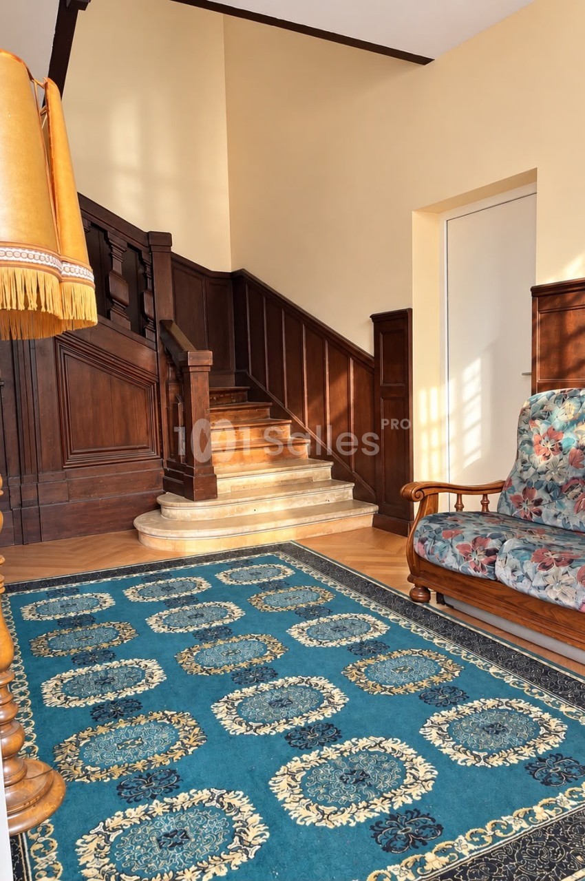 Escalier en pierre clair à double volée avec rampe en bois sombre, tapis bleu à motifs ornementaux et fauteuil fleuri dans un hall lumineux.