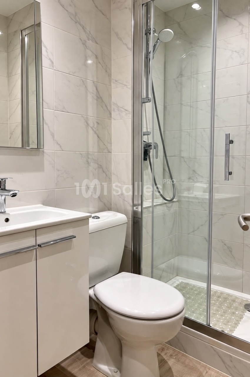 Salle de bain aux murs carrelés effet marbre, comprenant un lavabo sur meuble blanc, des toilettes et une douche à paroi vitrée.
