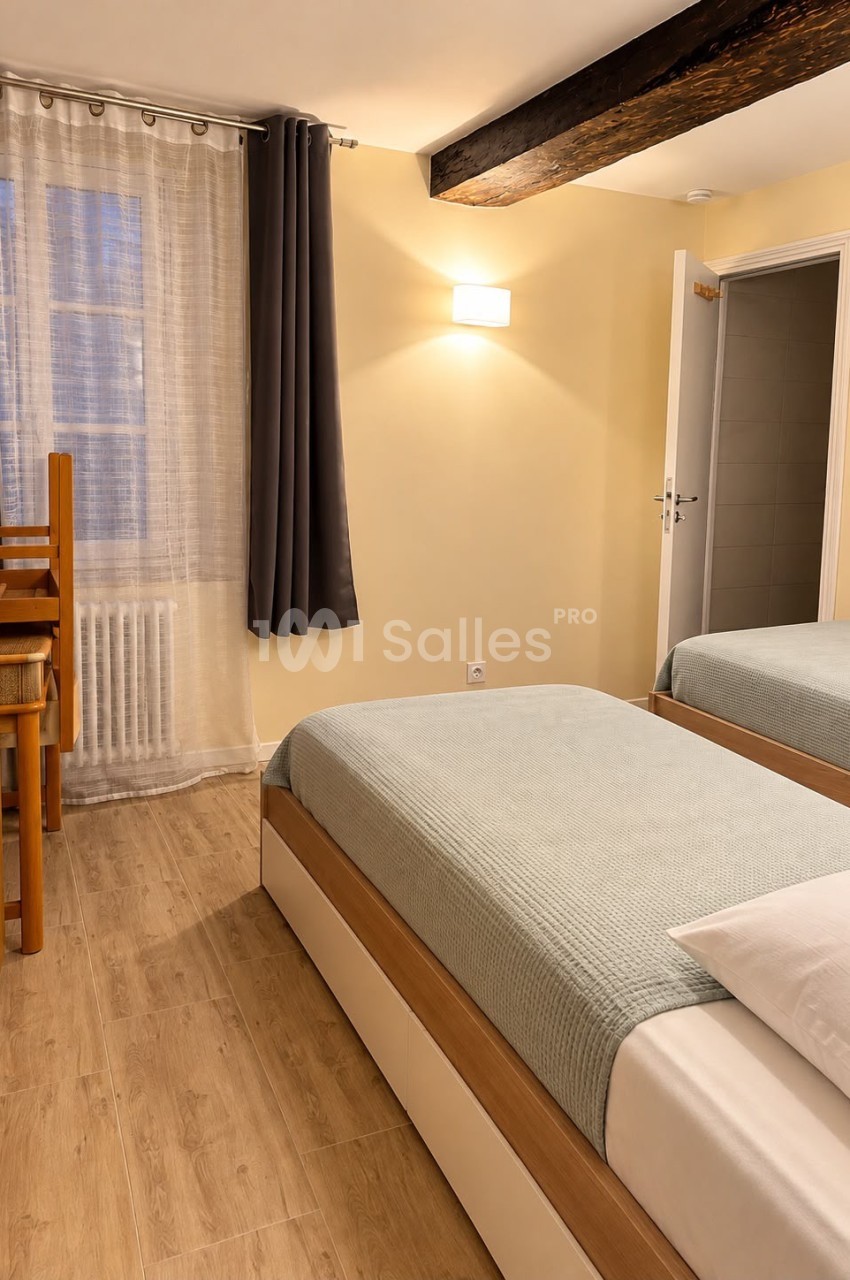 Chambre aux murs jaunes avec poutre apparente en bois sombre, deux lits simples recouverts de couvrelits vert d'eau, parquet clair et rideaux gris.