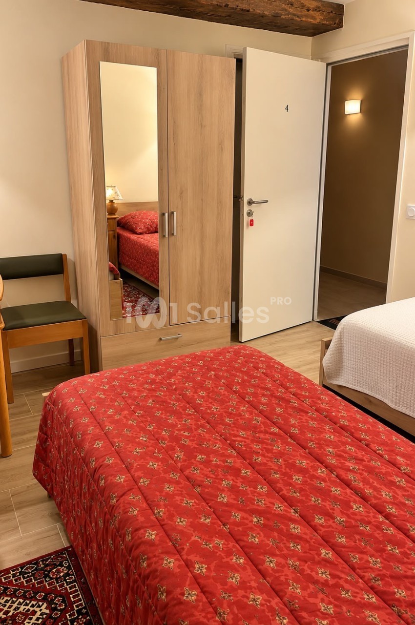 Chambre à coucher aux murs clairs avec un lit recouvert d'un couvre-lit rouge à motifs, une armoire en bois clair à miroir et une porte blanche ouverte.