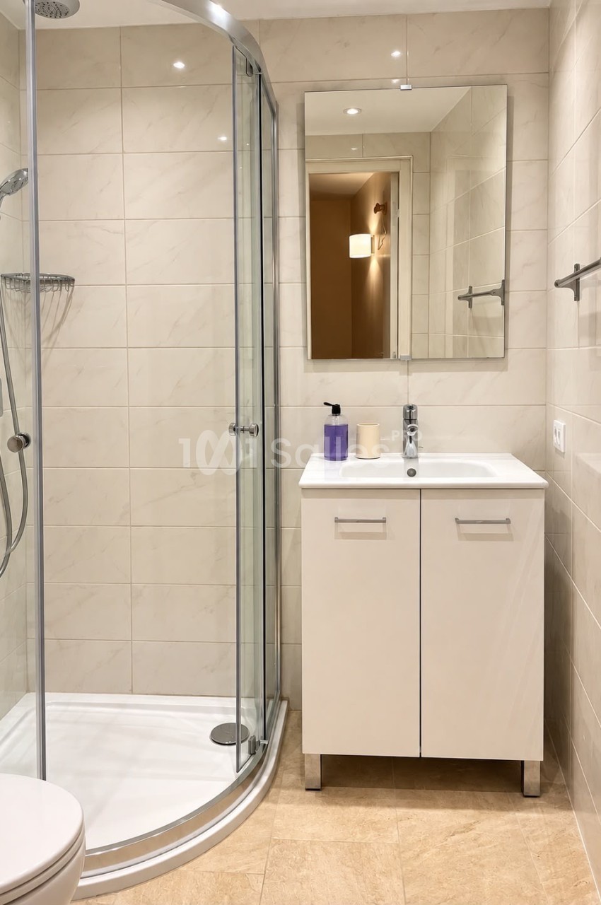 Salle de bain avec douche d'angle vitrée, vasque blanche sous un miroir et carrelage beige aux murs et au sol.