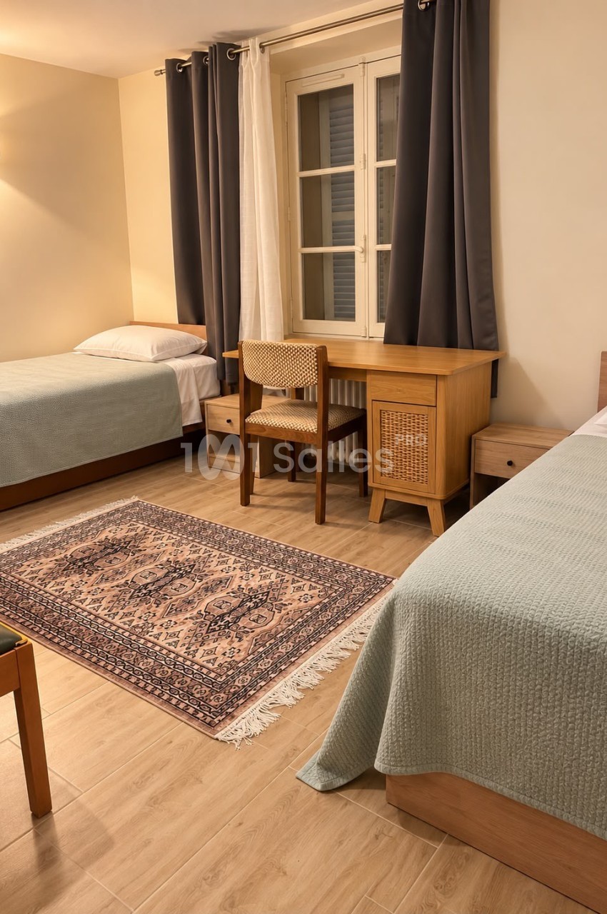 Chambre à deux lits individuels couverts de couvre-lits vert sauge, avec bureau en bois et chaise cannée près d'une fenêtre aux rideaux gris.