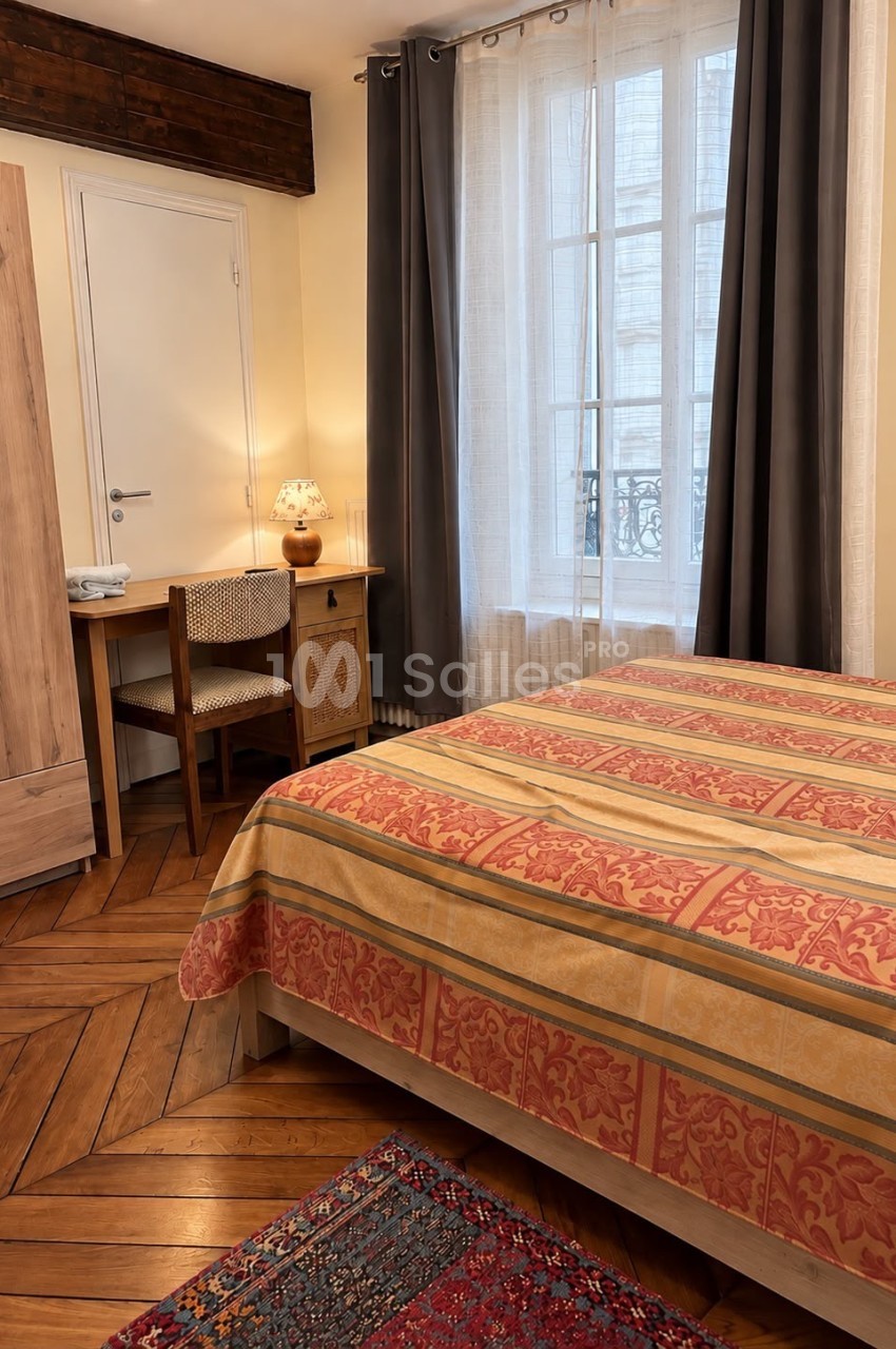 Chambre à coucher avec un lit double au couvre-lit rayé orange, un bureau en bois et une lampe, sur parquet en chevrons.