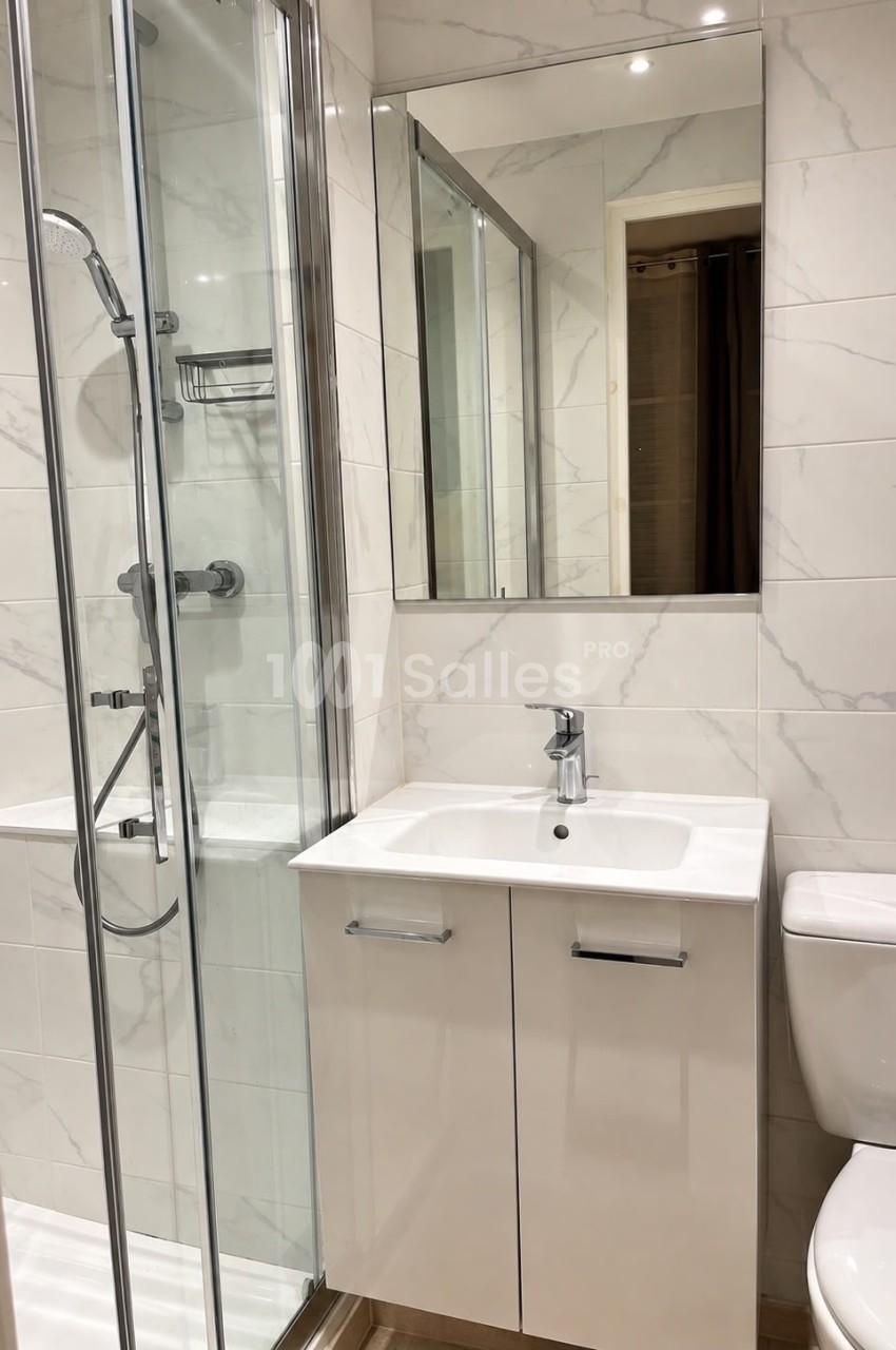 Salle de bains aux murs carrelés de marbre blanc, avec douche en cabine vitrée, lavabo blanc sur meuble et miroir, WC à droite.