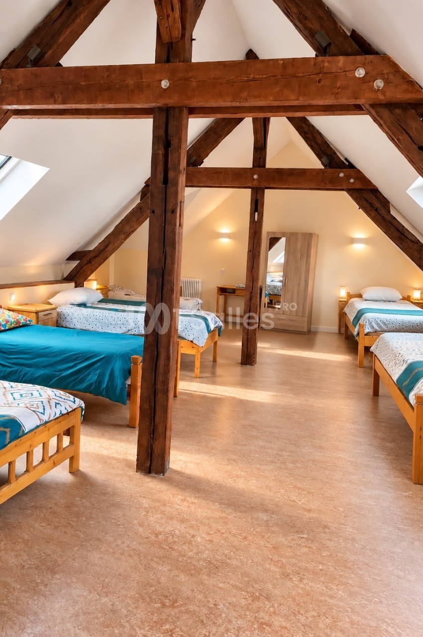 Dortoir sous combles aux poutres apparentes en bois, avec plusieurs lits simples aux couvertures bleu canard et une armoire en bois clair.