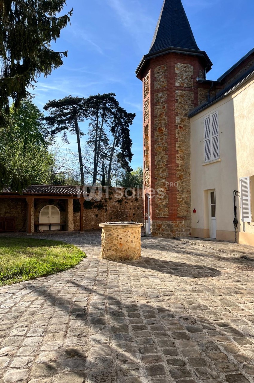 Cour pavée en pierre avec un puits central en pierre, face à une tour en brique et meulière surmontée d'un toit pointu, et un pavillon couvert en bois à gauche.