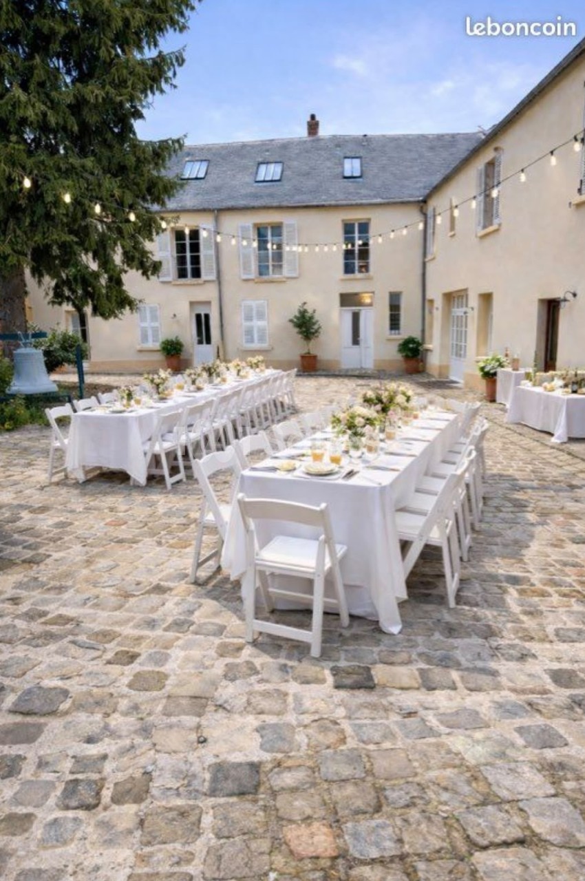 Cour pavée en extérieur dressée de longues tables nappées de blanc, chaises pliantes blanches et guirlandes lumineuses suspendues sur une façade de pierre claire. Let me check length: roughly 165 characters - too long. Need ≤125. Shorter: "Cour pavée