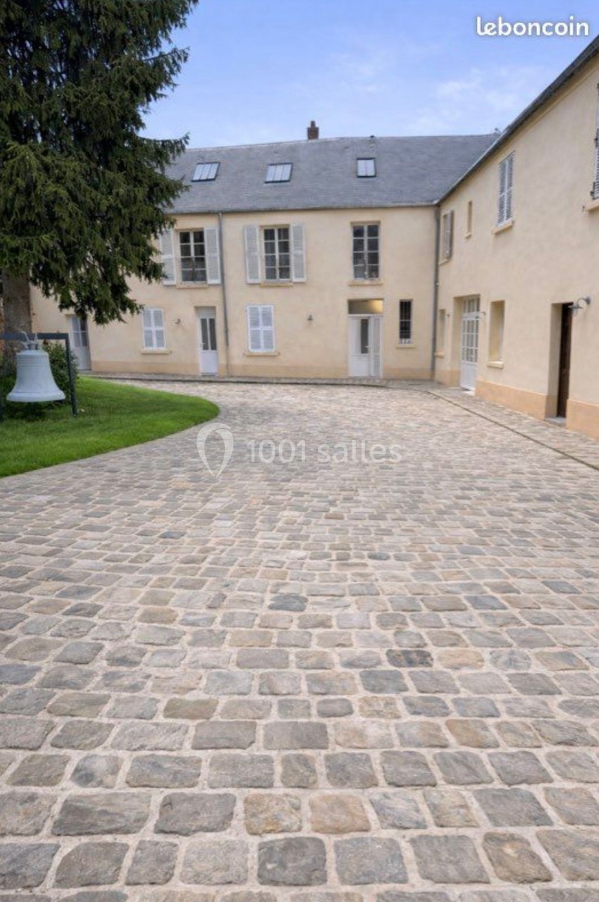 Cour pavée devant un bâtiment beige à deux étages avec fenêtres blanches, entouré d'arbres et de pelouse.