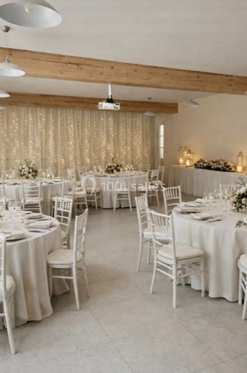Salle de réception décorée avec des tables rondes, nappes blanches, chaises assorties et guirlandes lumineuses en arrière…