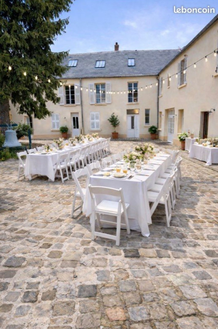 Tables dressées pour un repas en extérieur dans une cour pavée, entourée de bâtiments en pierre et décorée de guirlandes…