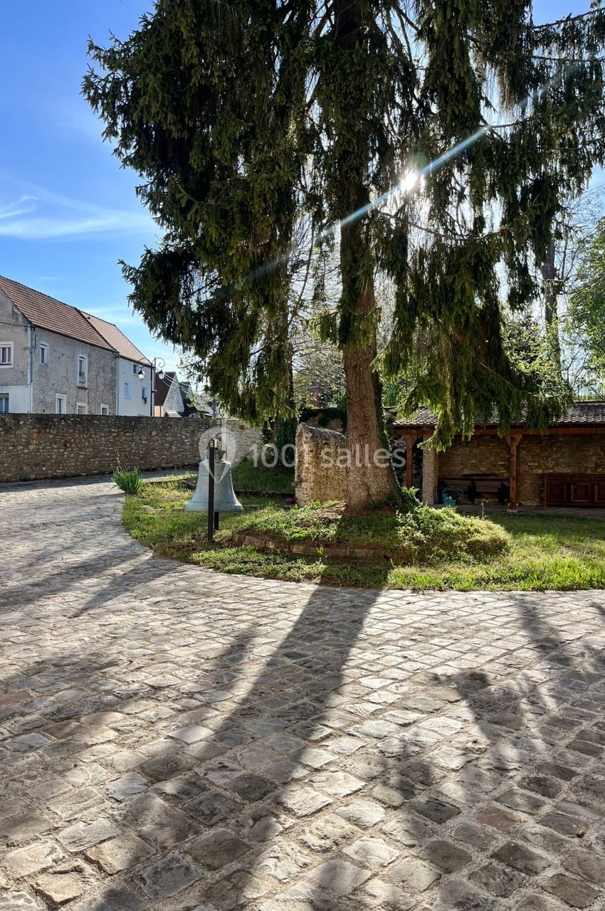 Cour pavée avec un grand sapin, une cloche en métal et un abri en bois, éclairés par un soleil matinal.