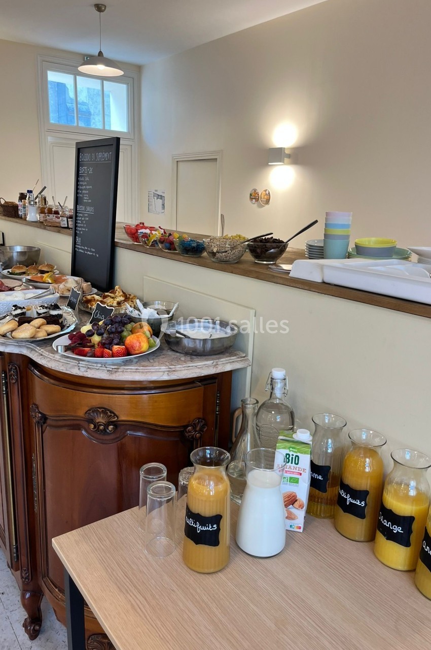 Buffet de petit-déjeuner avec viennoiseries, fruits, jus, lait et céréales disposés sur un meuble en bois.