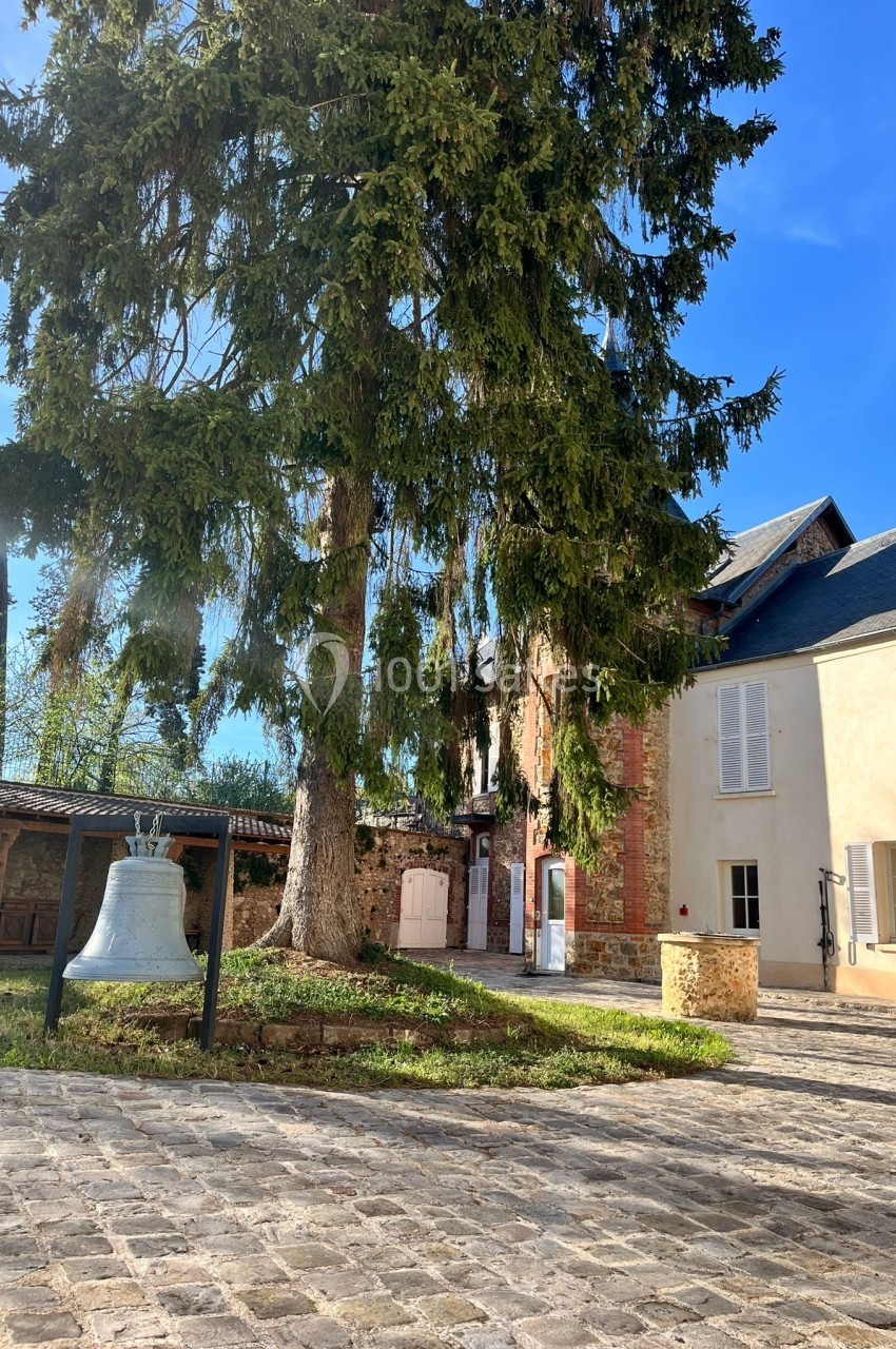 Cour pavée avec un grand sapin, une cloche exposée et des bâtiments anciens en arrière-plan sous un ciel bleu.