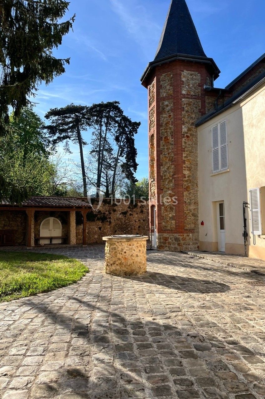 Cour pavée avec un puits central, une tour en pierre et des bâtiments aux volets blancs sous un ciel bleu.