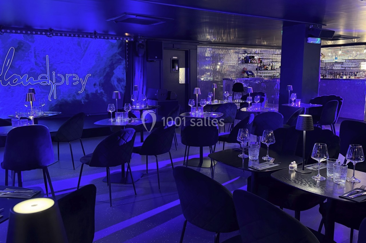 Salle de restaurant moderne avec éclairage bleu, tables dressées et un bar illuminé en arrière-plan.