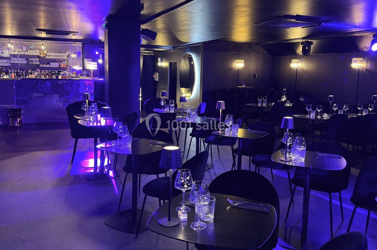 Salle de restaurant élégante avec éclairage tamisé violet, tables dressées et bar en arrière-plan.