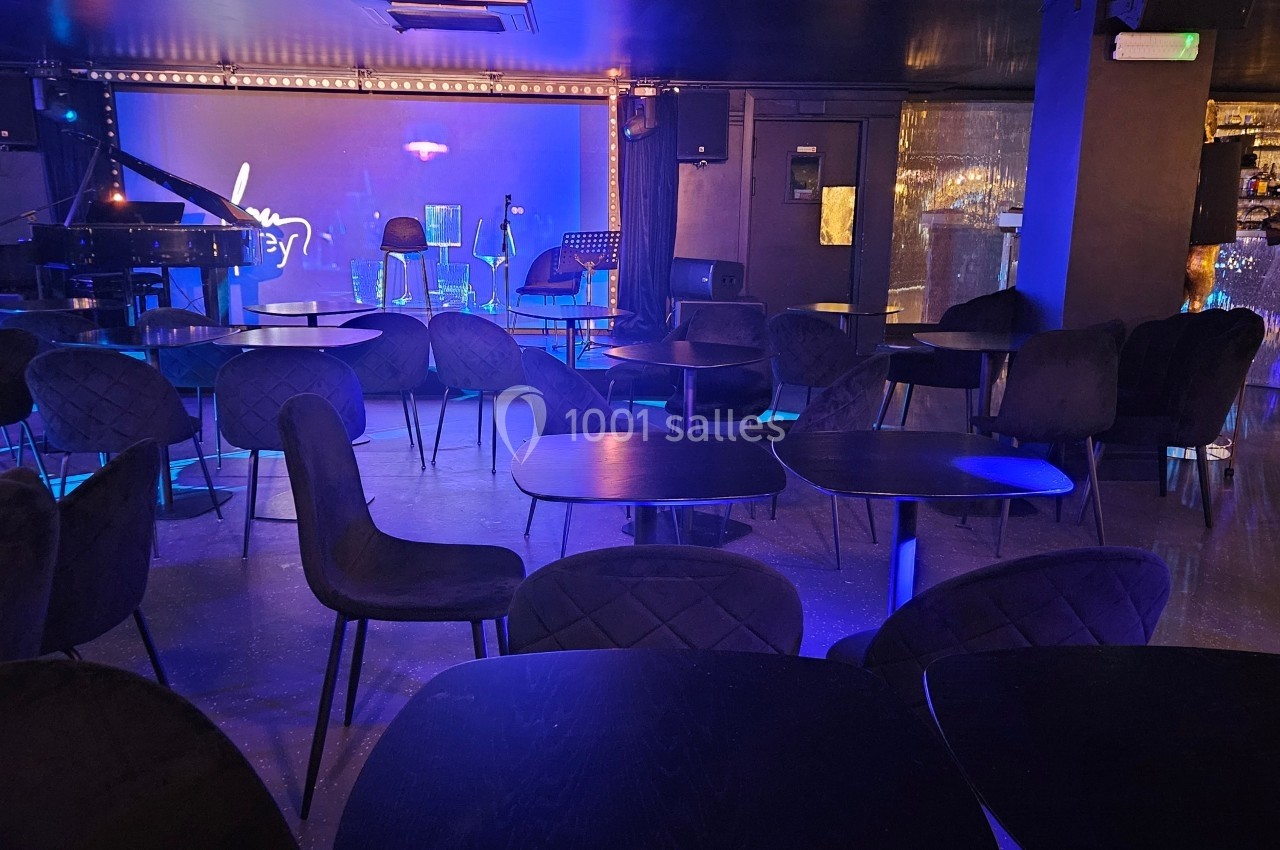 Salle de spectacle avec tables et chaises vides, éclairée par une lumière bleue, scène équipée d'un piano et de micros.
