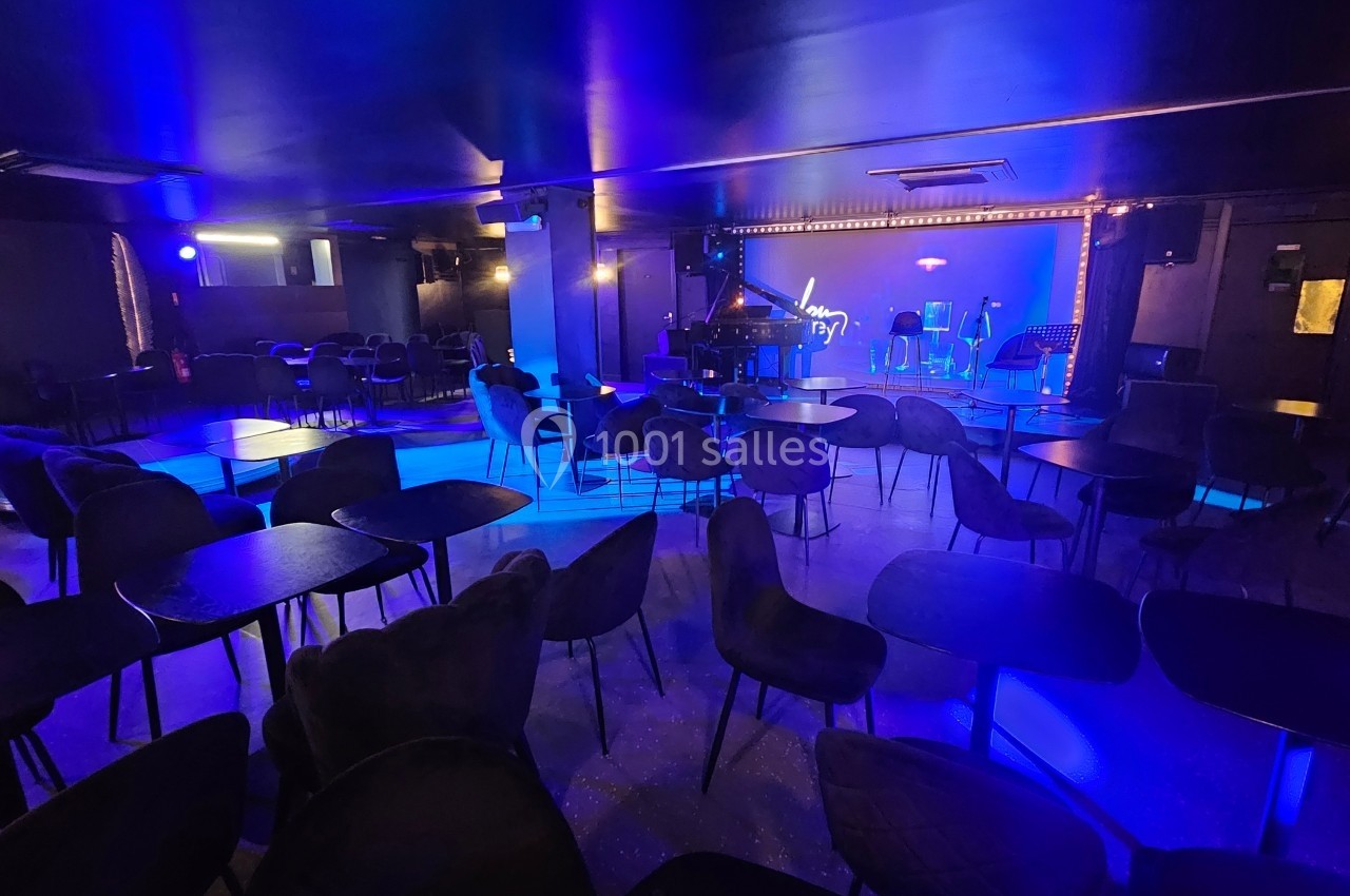 Salle de spectacle avec tables et chaises disposées face à une scène éclairée en bleu, équipée d'un piano et de micros.