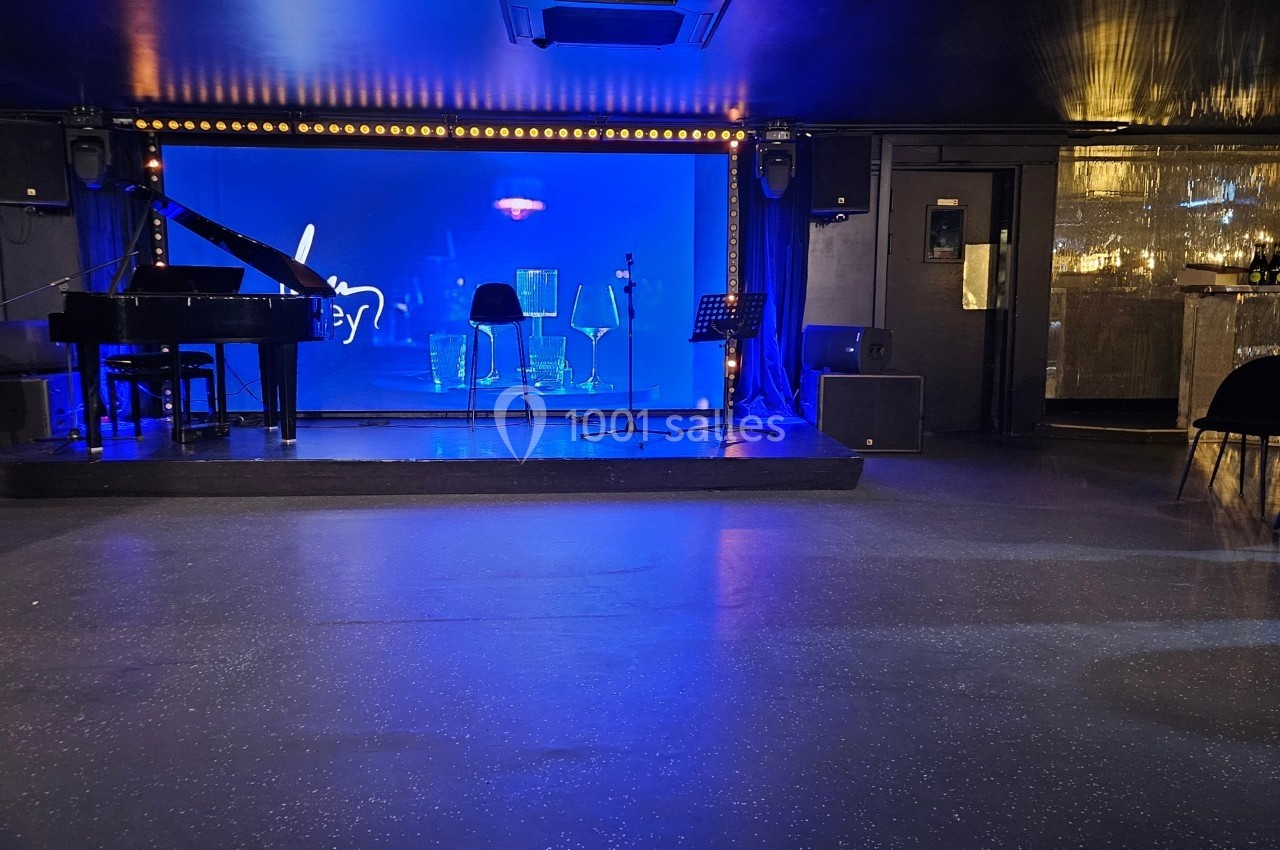 Scène vide avec un piano à queue, deux tabourets et des micros, éclairée par une lumière bleue dans une salle sombre.