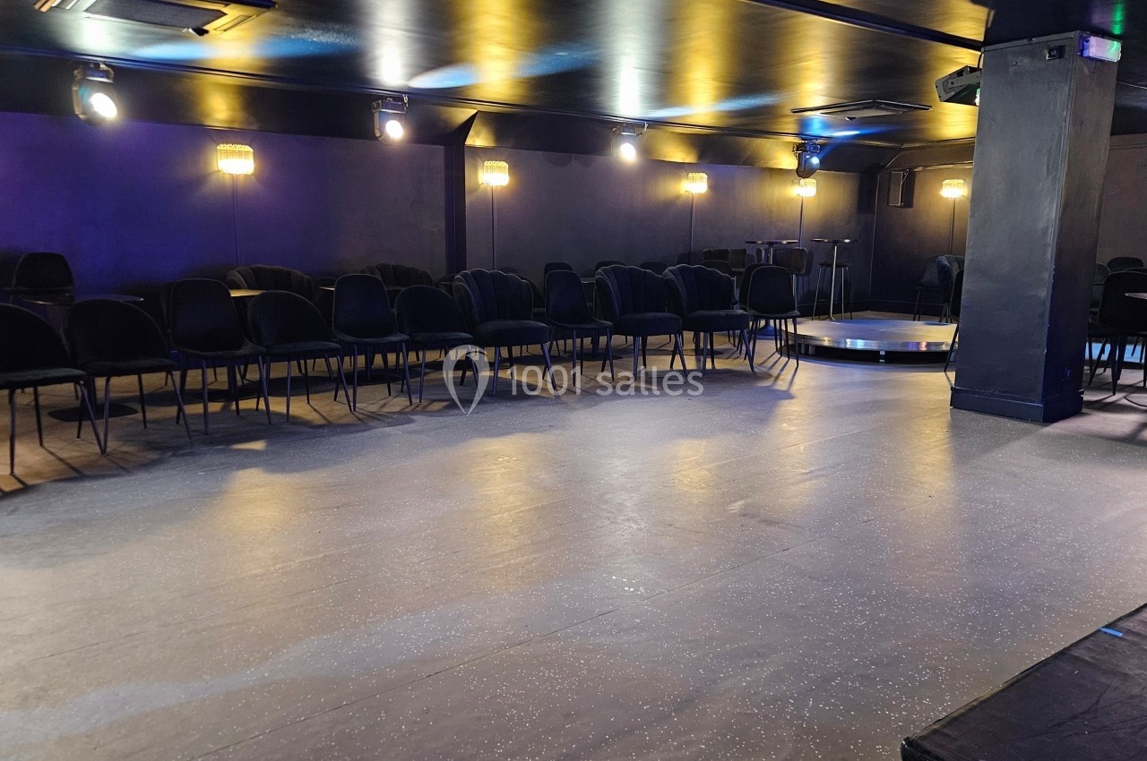 Salle sombre avec des rangées de chaises noires disposées face à une petite scène circulaire éclairée.