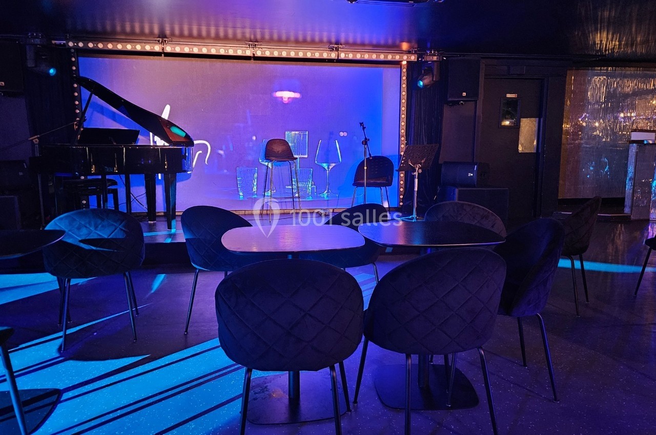 Salle de spectacle avec scène éclairée en bleu, équipée d'un piano, de micros et de chaises devant des tables.