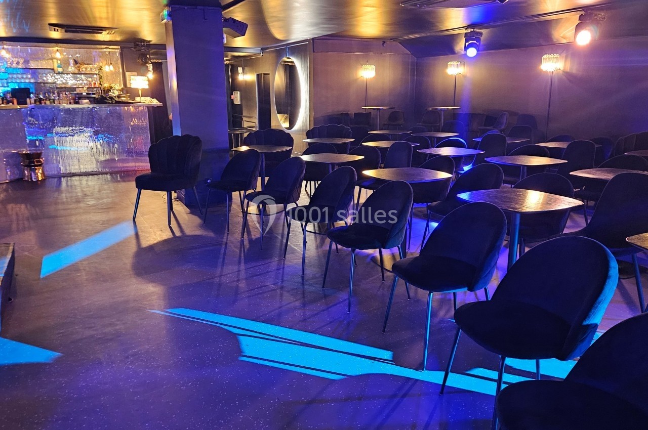 Salle avec des tables et chaises noires, éclairée par une lumière bleue, avec un bar en arrière-plan.