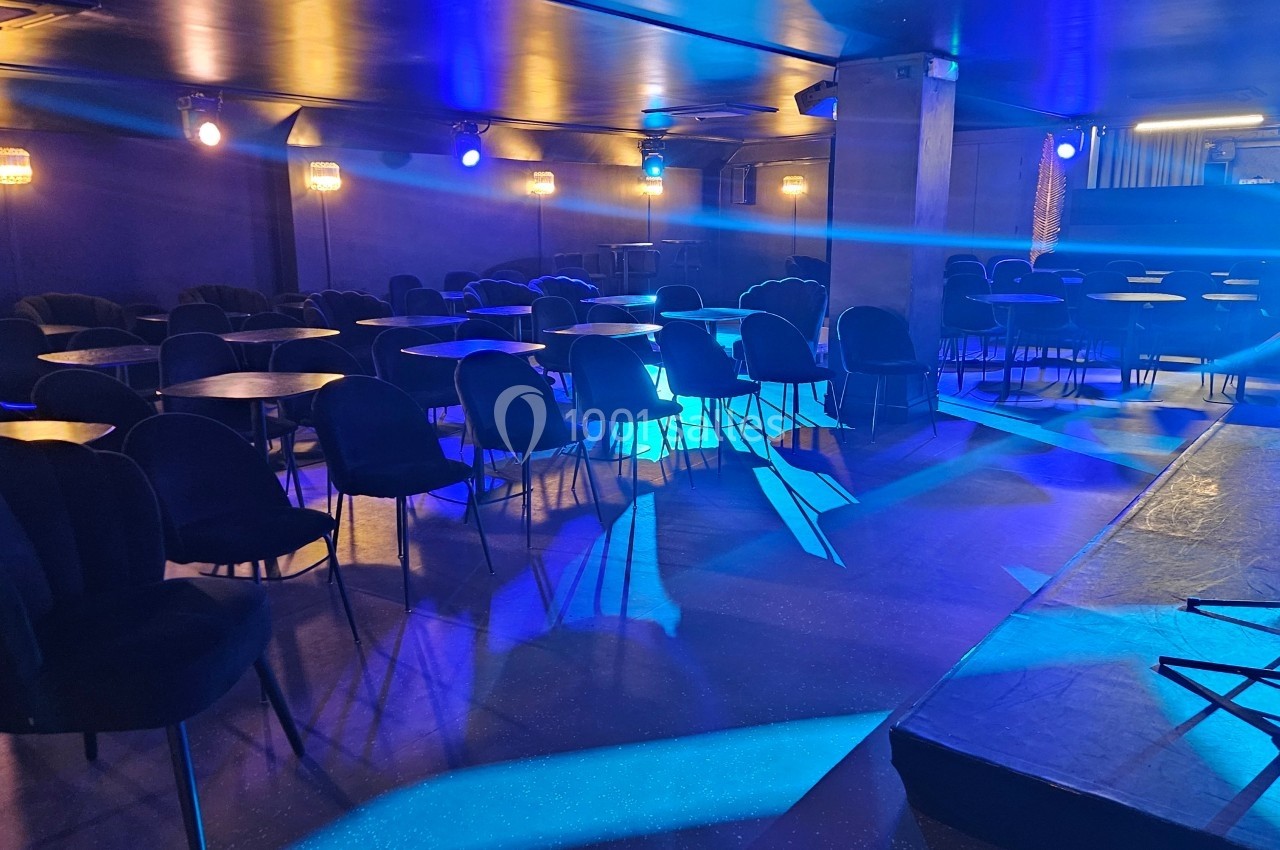 Salle sombre avec des tables rondes, des chaises noires et un éclairage bleu, vue depuis une scène.