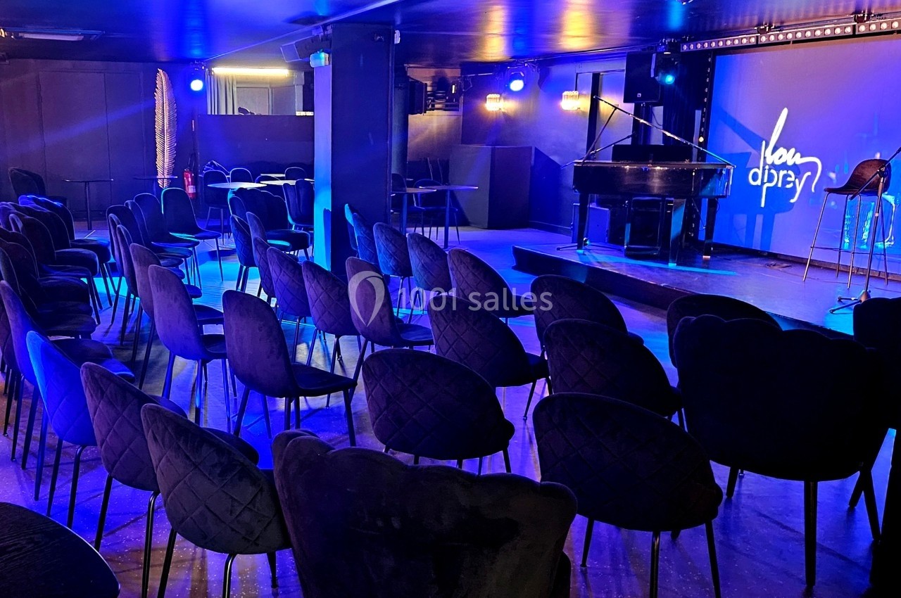 Salle de spectacle avec sièges alignés, éclairage bleu et scène équipée d'un piano à queue et d'un micro.