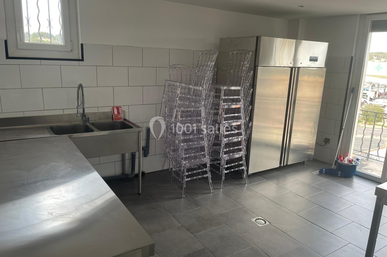 Cuisine professionnelle avec évier, plan de travail en inox, réfrigérateur double porte et chaises empilées.