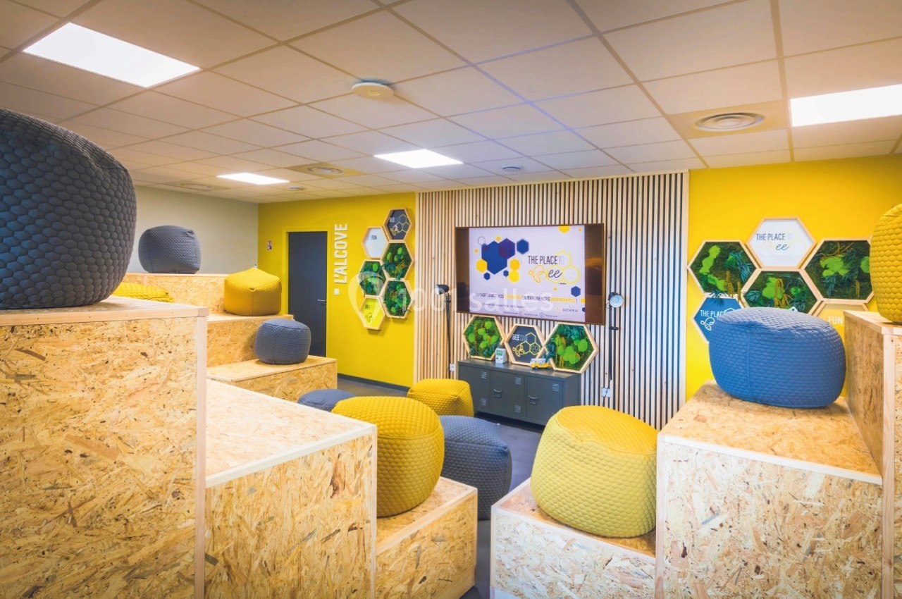 Salle colorée avec poufs et gradins en bois, décorée de panneaux hexagonaux et d'un écran mural.