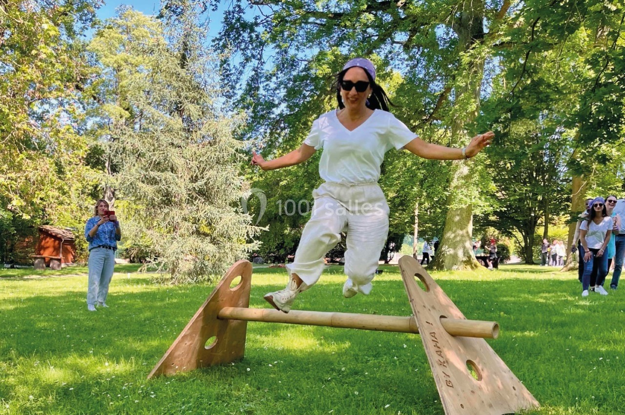 Une femme saute par-dessus une barre en bois dans un parc verdoyant, sous un ciel ensoleillé.