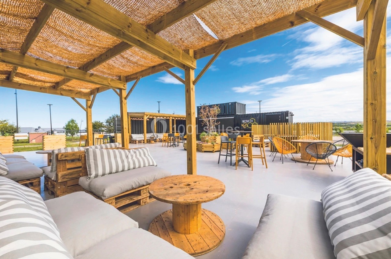 Terrasse extérieure avec pergola en bois, mobilier en palettes et vue dégagée sur un ciel partiellement nuageux.