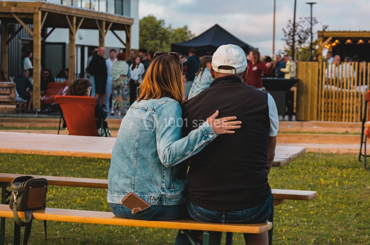 Un couple assis sur un banc en bois regarde un rassemblement en plein air dans une ambiance conviviale.