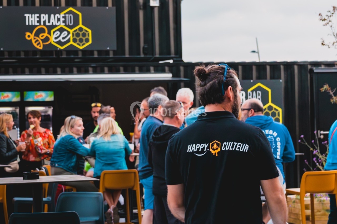 Un groupe de personnes discutant devant un bar en extérieur, avec un homme portant un t-shirt ’Happy Culteur’ au premier…