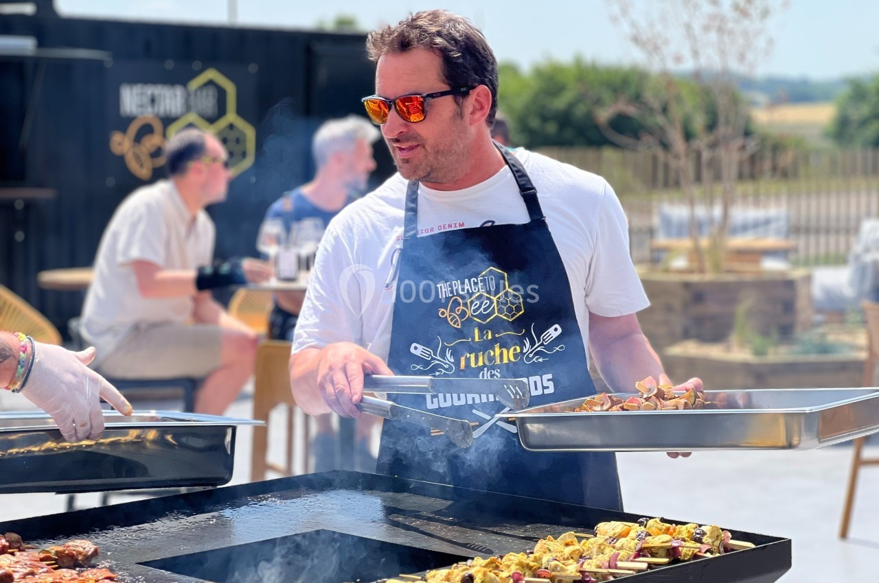 Un homme en tablier cuisine des brochettes sur une plancha en extérieur, avec des personnes en arrière-plan.