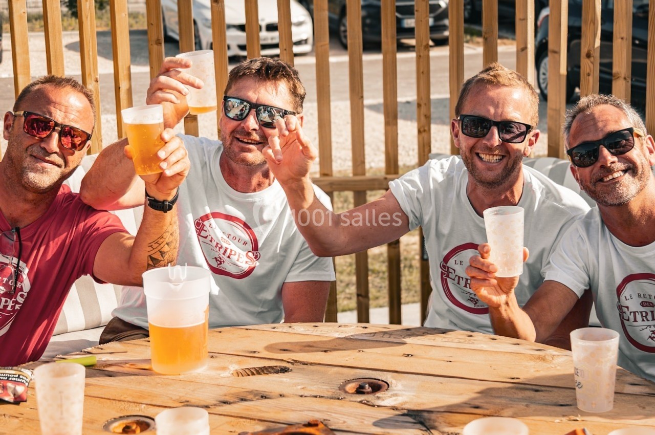 Quatre hommes souriants assis autour d'une table en bois, levant leurs verres en extérieur par temps ensoleillé.