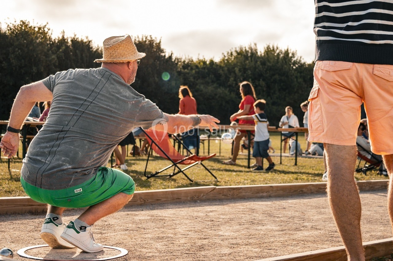 Un homme lance une boule de pétanque sur un terrain en plein air, entouré de spectateurs et de joueurs.
