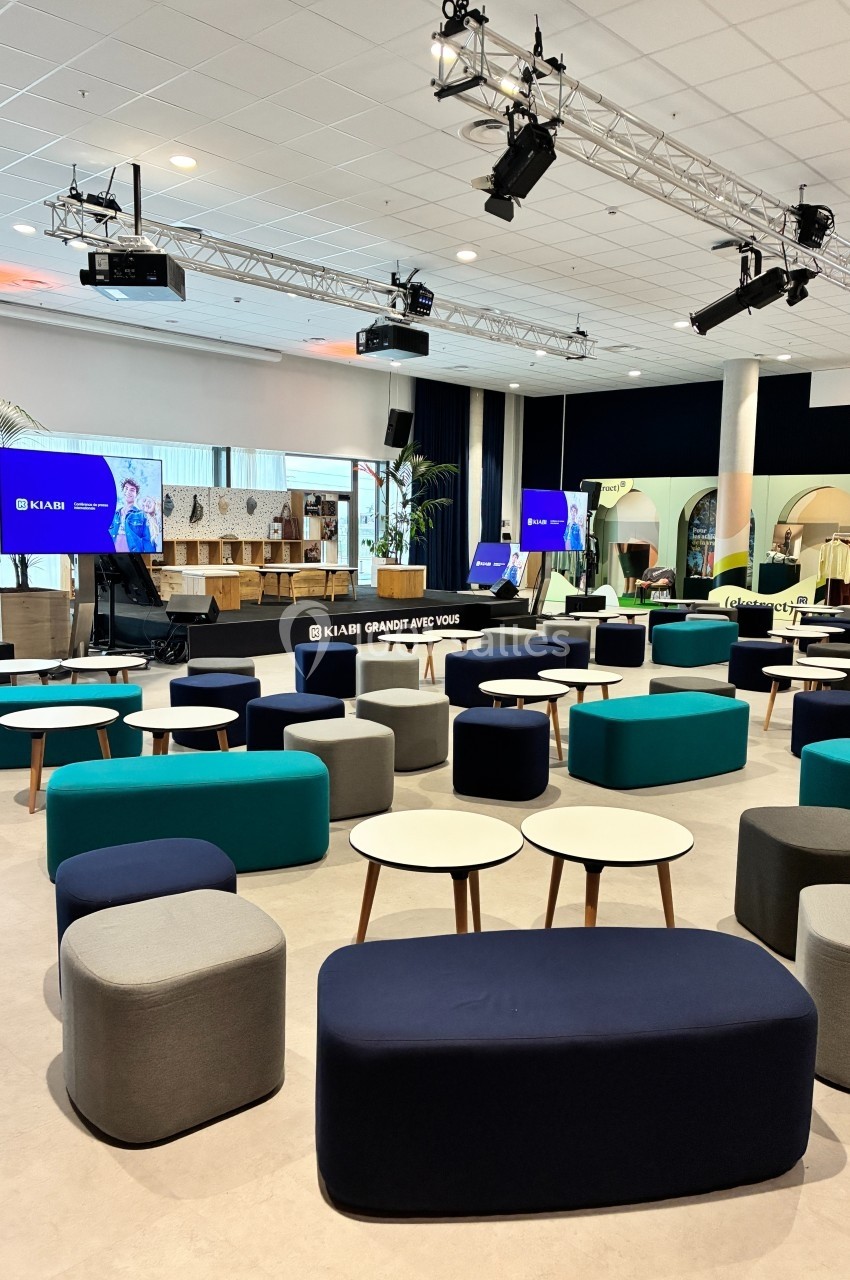 Salle de conférence moderne avec poufs colorés, tables basses et écrans affichant une présentation.