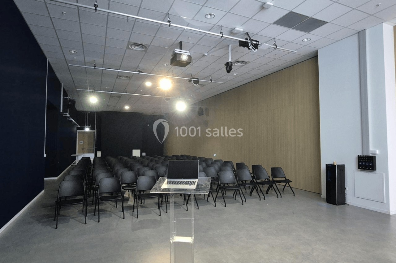 Salle de conférence vide avec des rangées de chaises noires, un pupitre transparent et des projecteurs allumés.