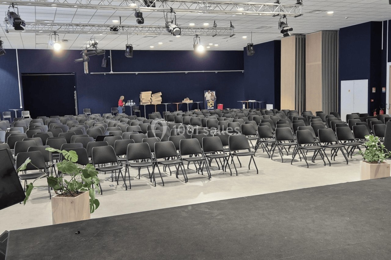 Salle de conférence avec des rangées de chaises noires alignées, vue depuis une scène équipée d'écrans et de plantes.