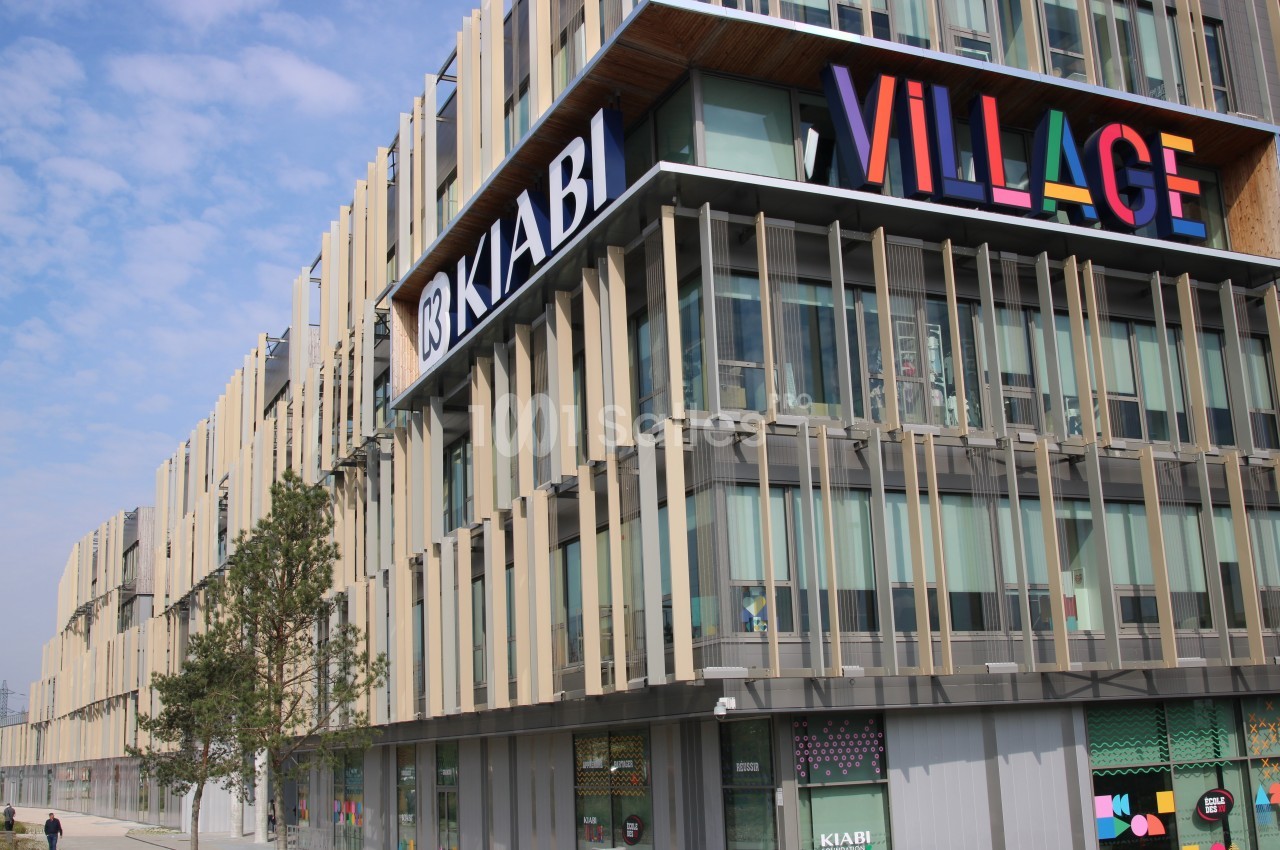 Façade moderne d'un centre commercial avec enseignes colorées, dont ’Kiabi’ et ’Village’, sous un ciel partiellement nuageux.