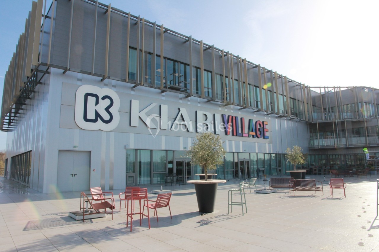 Façade d'un bâtiment moderne avec le logo ’Kiabi Village’, entourée de tables et chaises sur une terrasse ensoleillée.