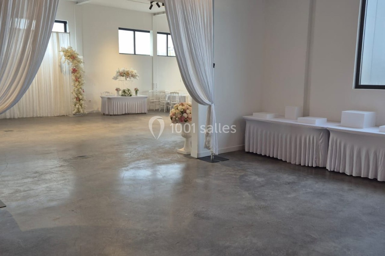Salle lumineuse avec sol en béton, rideaux blancs, tables drapées et décorations florales en arrière-plan.