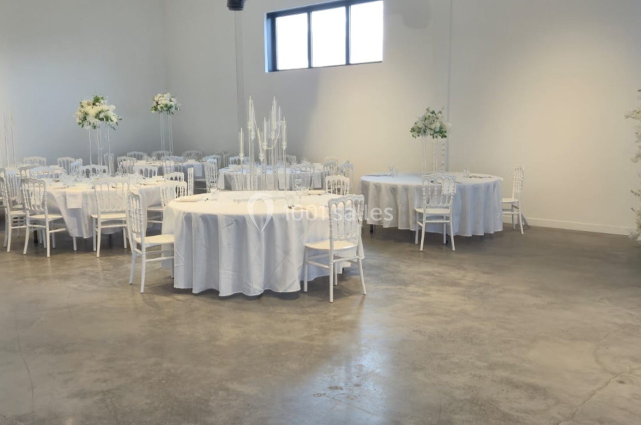 Salle de réception lumineuse avec tables rondes dressées, chaises blanches et décorations florales élégantes.