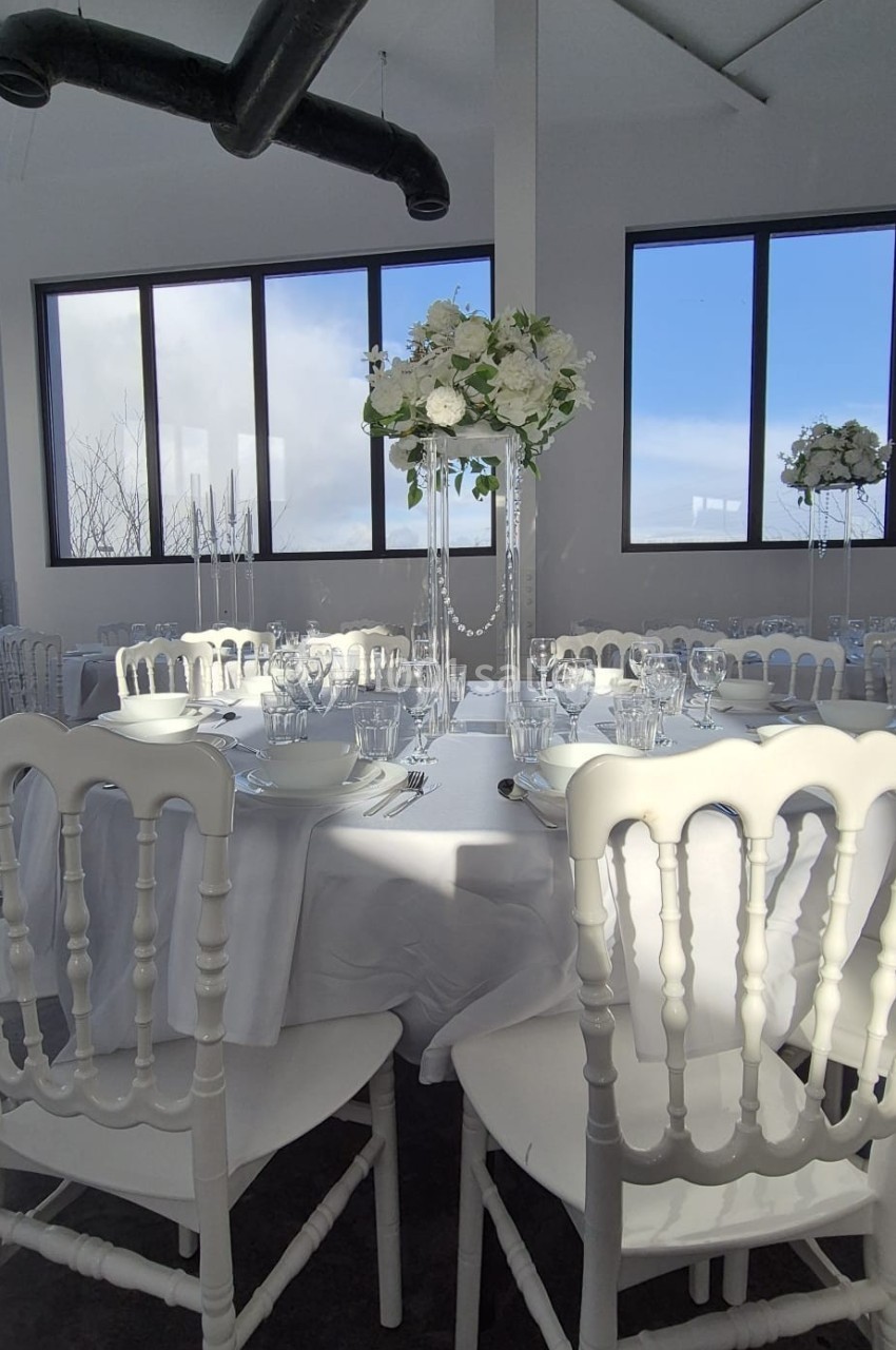 Salle lumineuse avec table ronde dressée, chaises blanches et centre de table floral sous de grandes fenêtres.