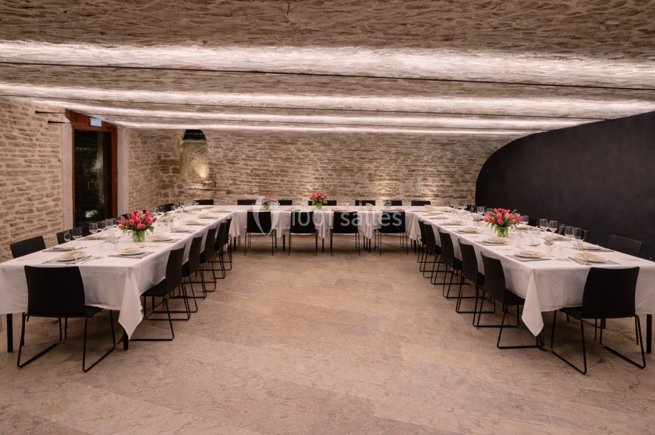 Salle de réception avec tables dressées en U, nappes blanches, chaises noires et bouquets de fleurs roses au centre.