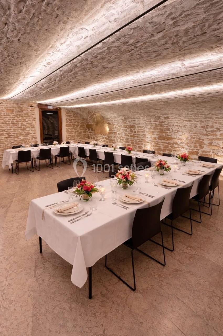 Salle voûtée en pierre avec tables dressées, nappes blanches, chaises noires et décorations florales.