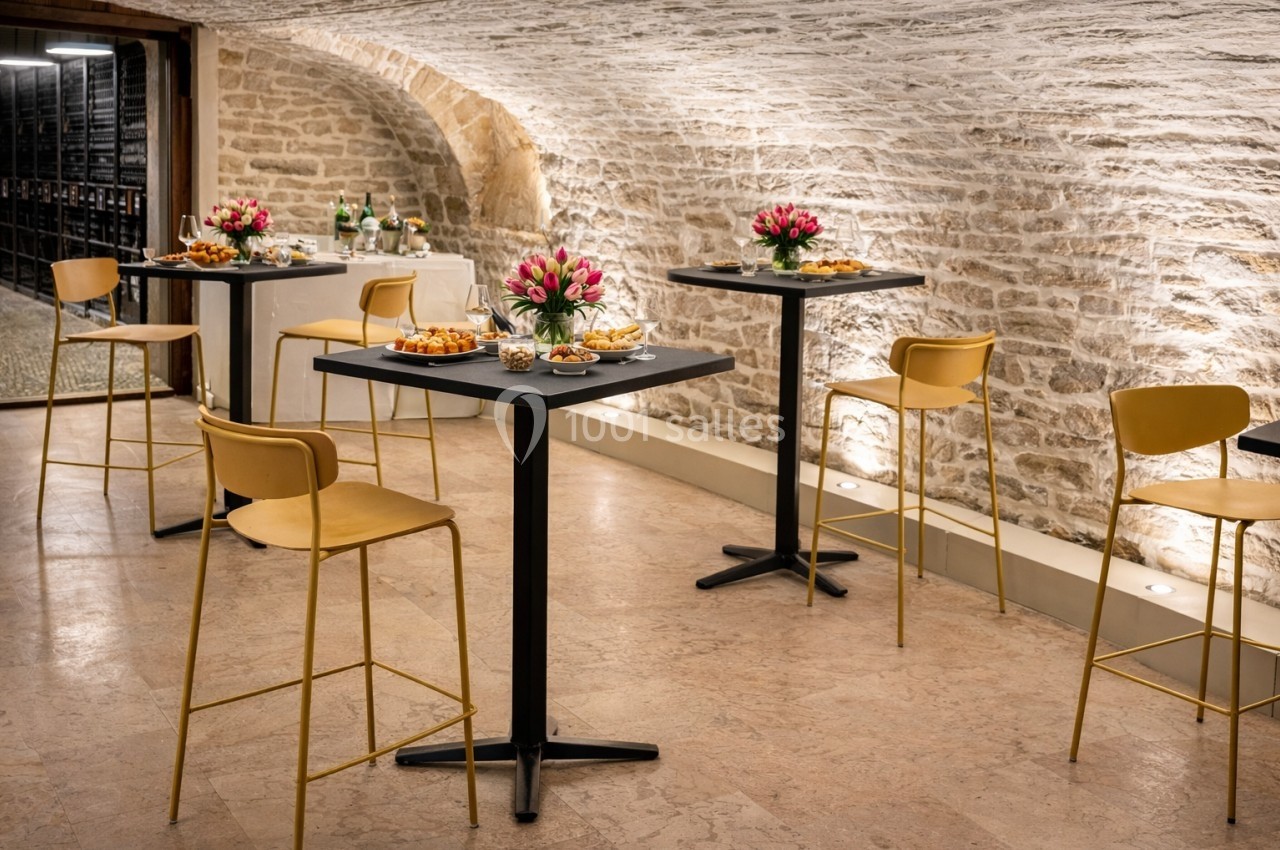 Salle voûtée en pierre avec tables hautes, chaises dorées et décorations florales, préparée pour un événement.