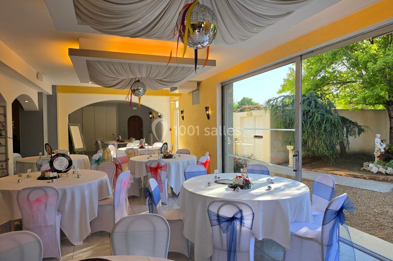 Salle de réception décorée avec des tables rondes, nappes blanches et chaises ornées de rubans colorés, vue sur un jardin.