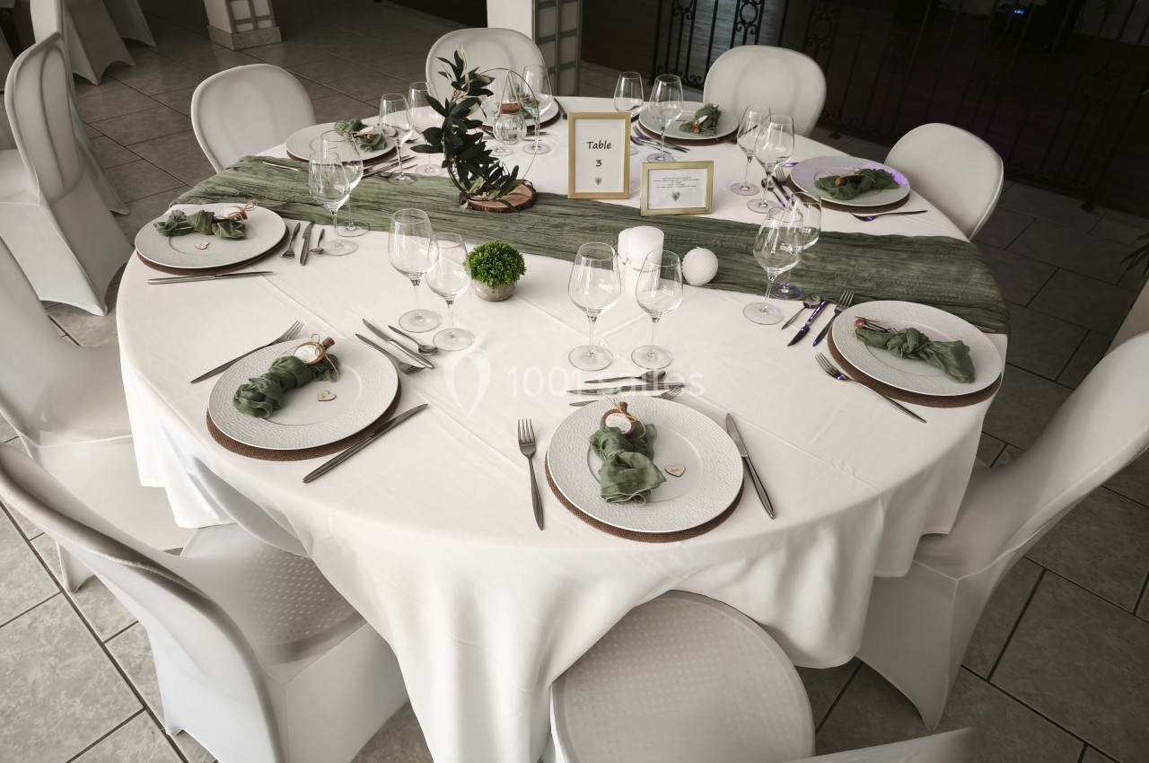 Table ronde élégamment dressée avec nappes blanches, serviettes vertes pliées et décoration végétale centrale.